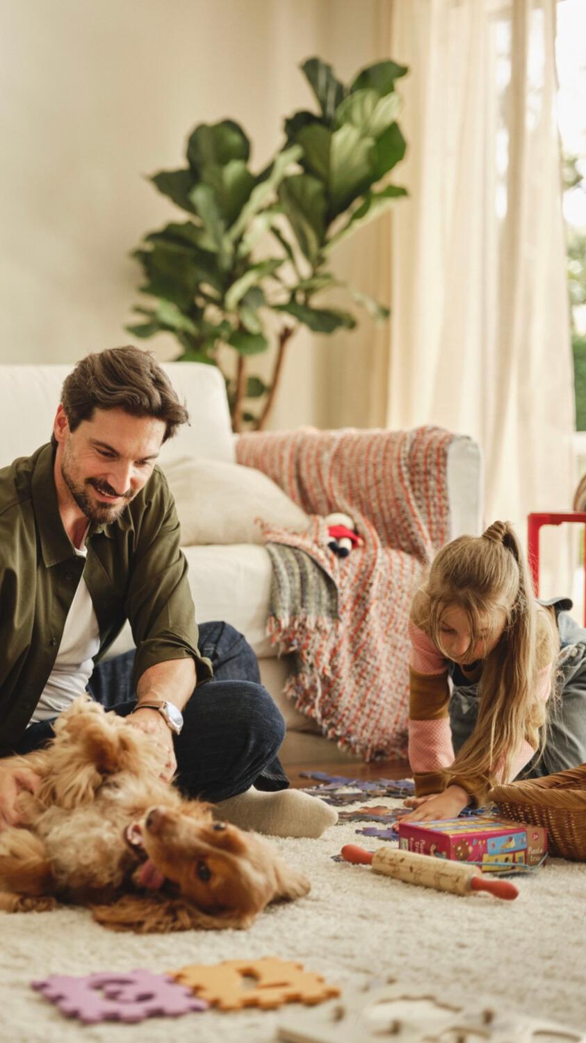 Man plays with dog while child does puzzles and woman reads on sofa.