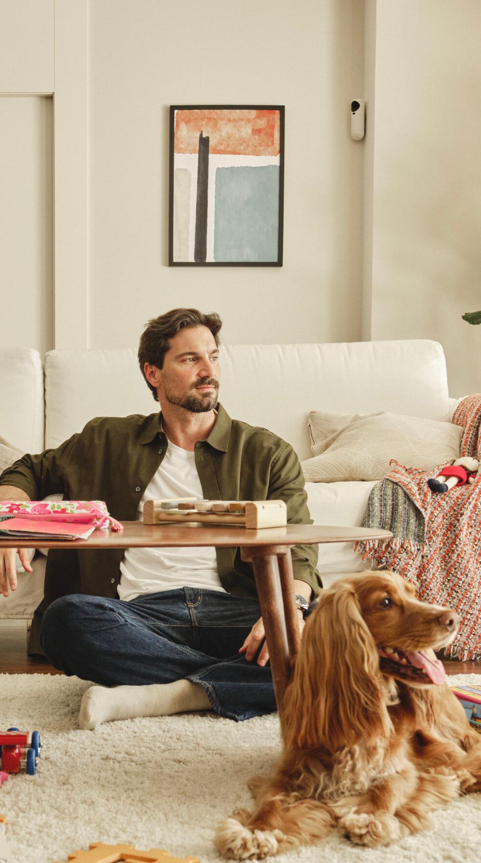 Man sitting on the floor in a living room next to a small table, with a dog lying in the foreground.